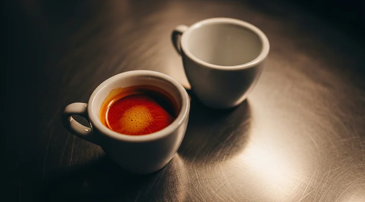 Primer plano de una taza blanca llena de café recién hecho sobre acero oscuro junto a una segunda taza idéntica todavía completamente vacía y desenfocada, representando dos clientes dados de alta el mismo día con resultados distintos